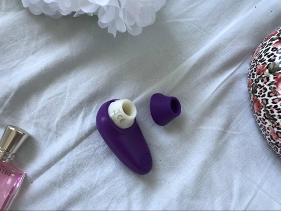 фиолетовый Womanizer Starlet лежит на белой кровати рядом с духами и косметичкой